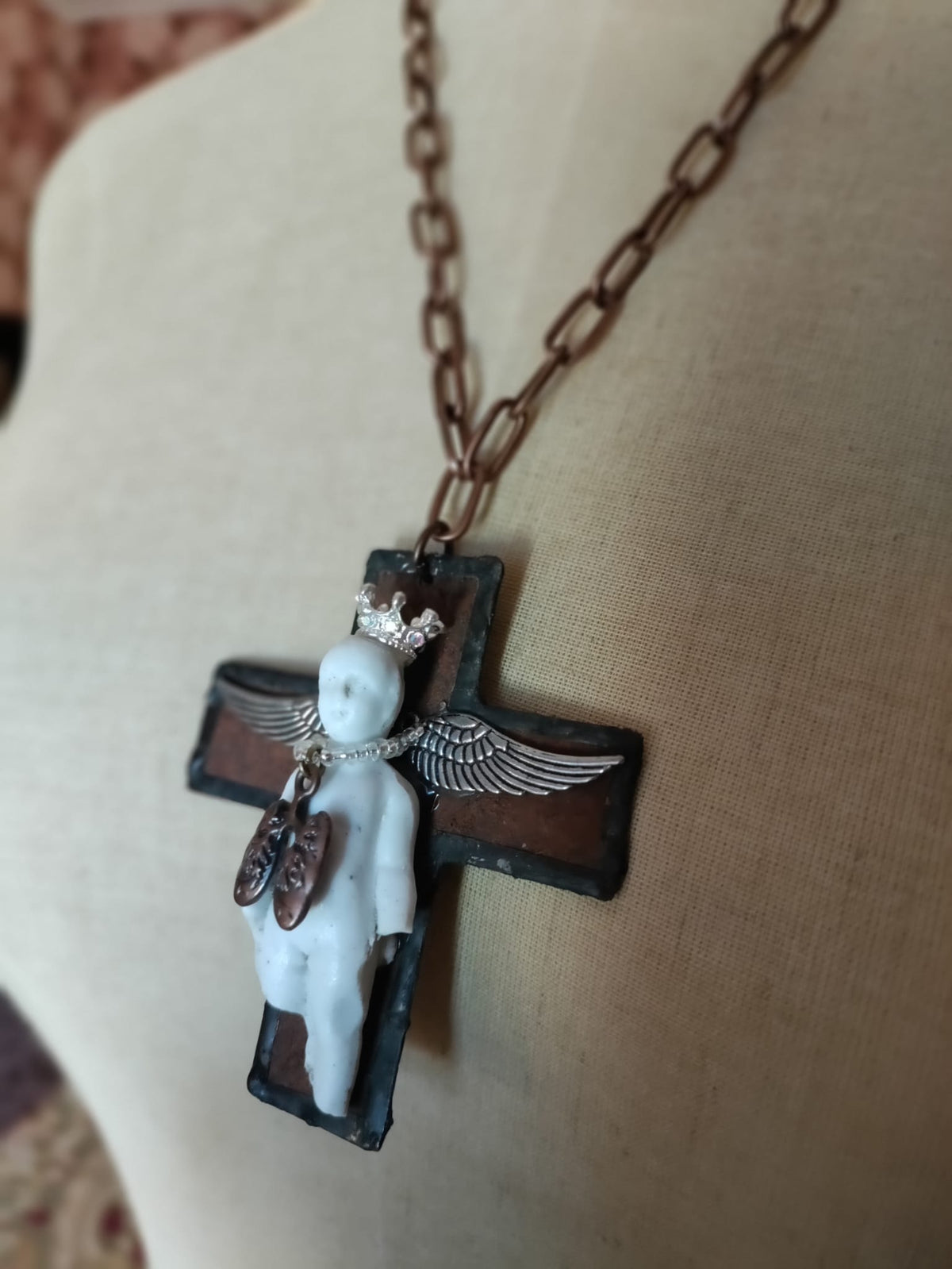 Angled view of Charles' Last Breath pendant, bisque doll on black cross with silver wings and copper lungs charm catching the light