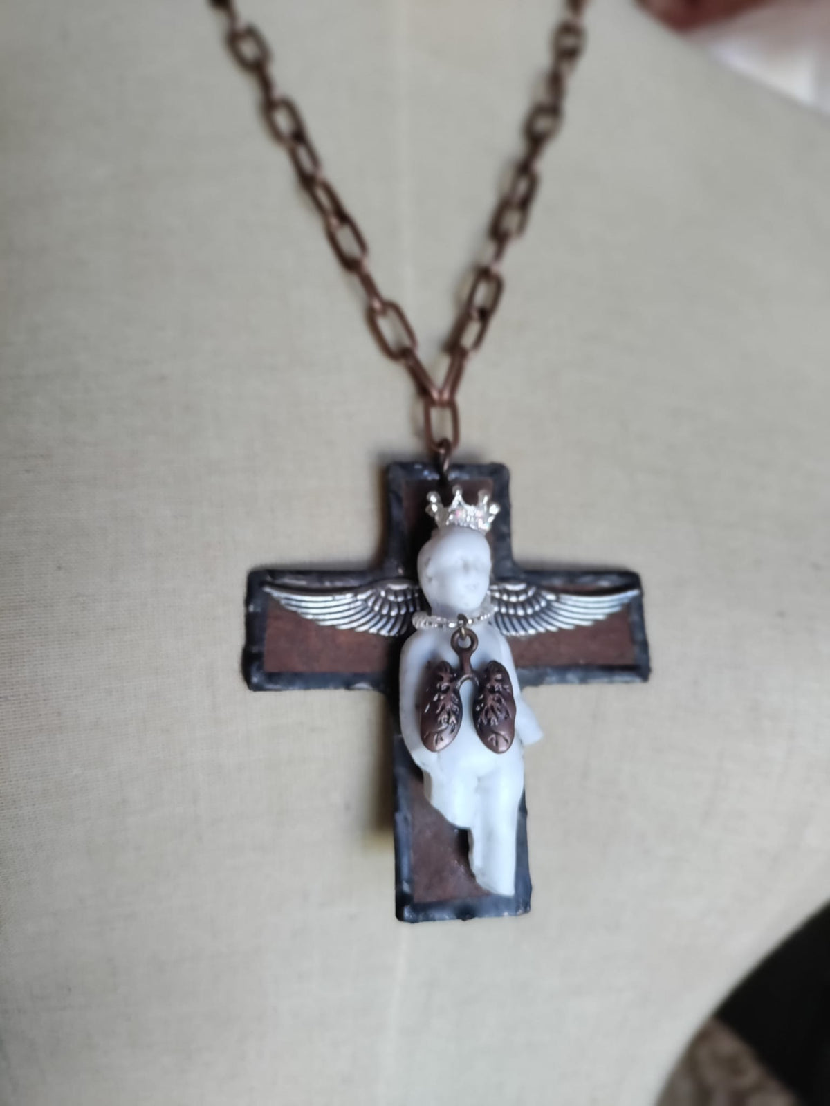 Charles' Last Breath necklace on mannequin, antique bisque doll on a black cross wearing a rhinestone crown and silver angel wings with a copper anatomical lungs charm