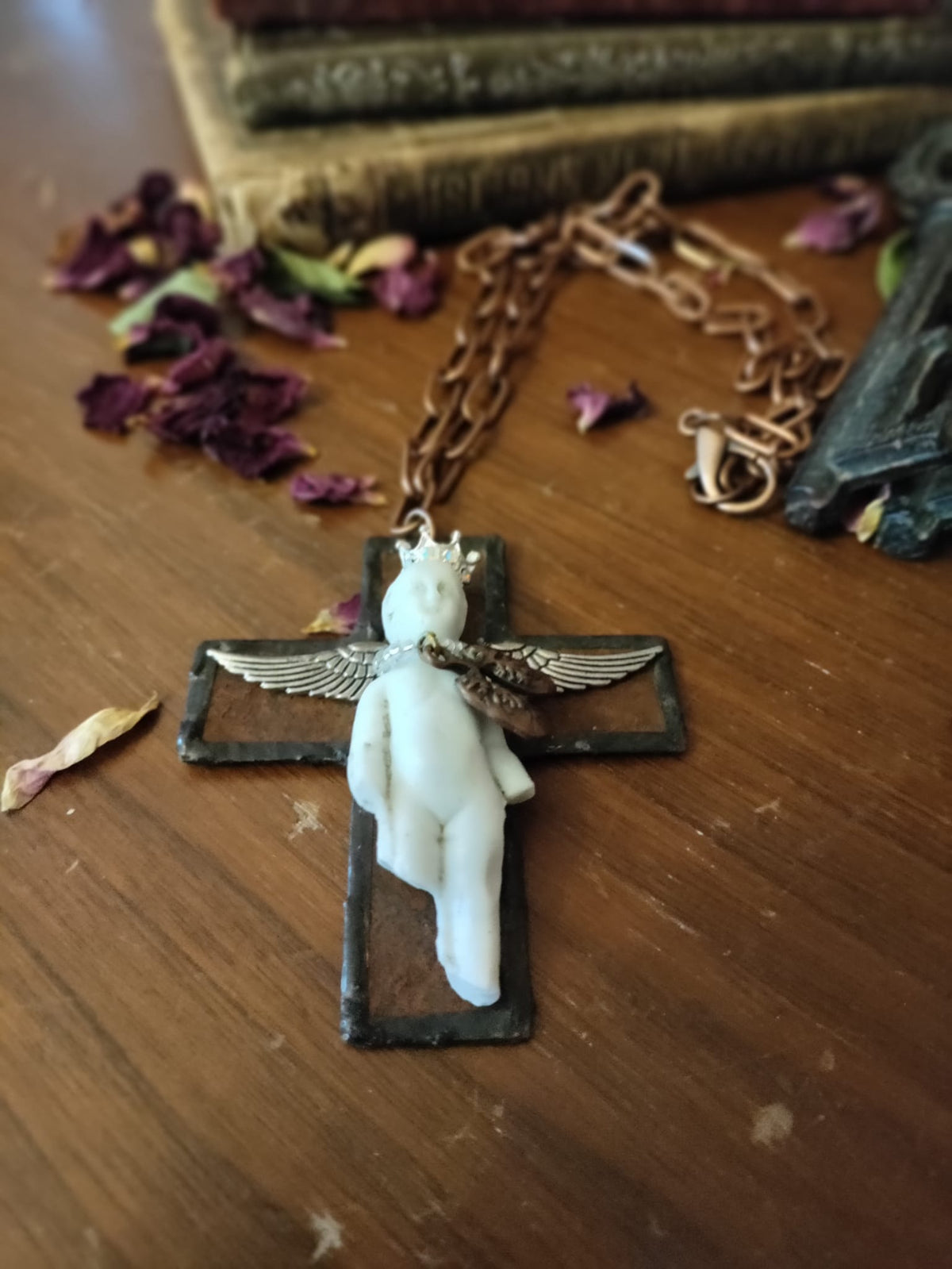 Close-up flat lay of Charles' Last Breath cross pendant with copper chain, dried rose petals, and antique books in the background