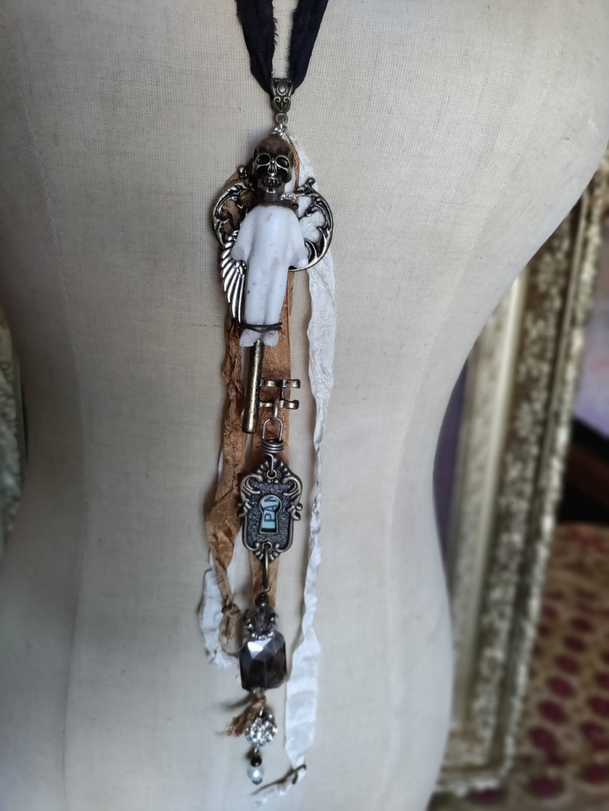 Skeleton Key Frozen Charles necklace on mannequin, full pendant showing bronze skull, silver angel wing, brass skeleton key, keyhole escutcheon charm, and faceted crystal drop on black silk ribbon