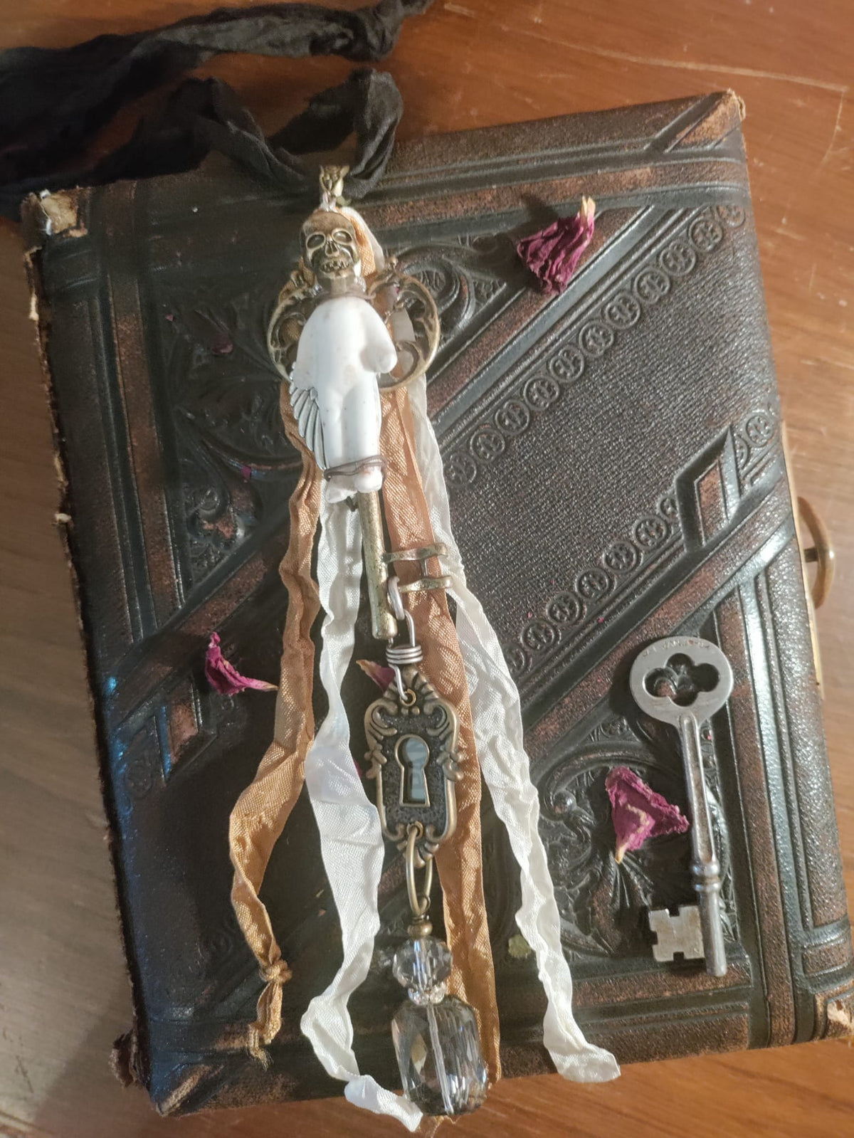 Skeleton Key Frozen Charles necklace flat lay on an antique book, bisque doll pendant with brass skeleton key, keyhole escutcheon charm, and faceted crystal drop beside a vintage skeleton key and dried rose petals