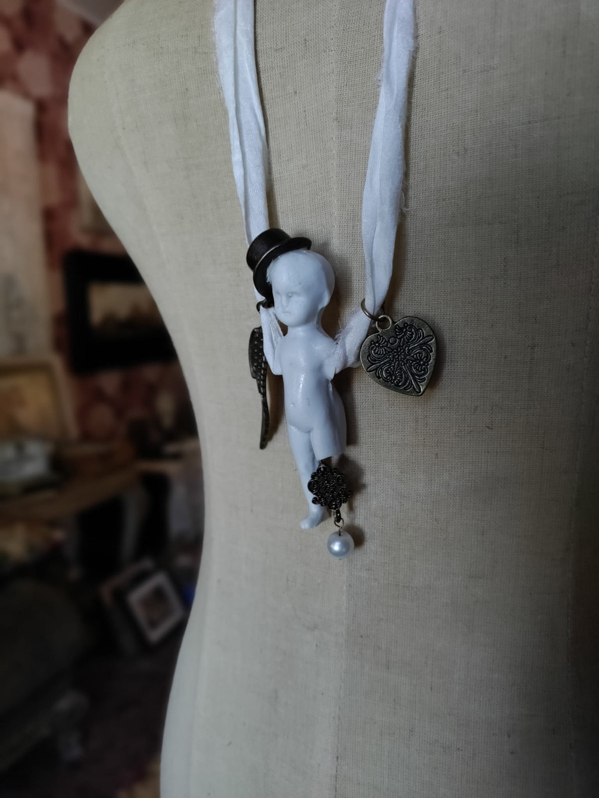 Cypher in the Snow necklace on mannequin, bisque doll pendant with bronze wing and heart charms against ivory silk ribbon