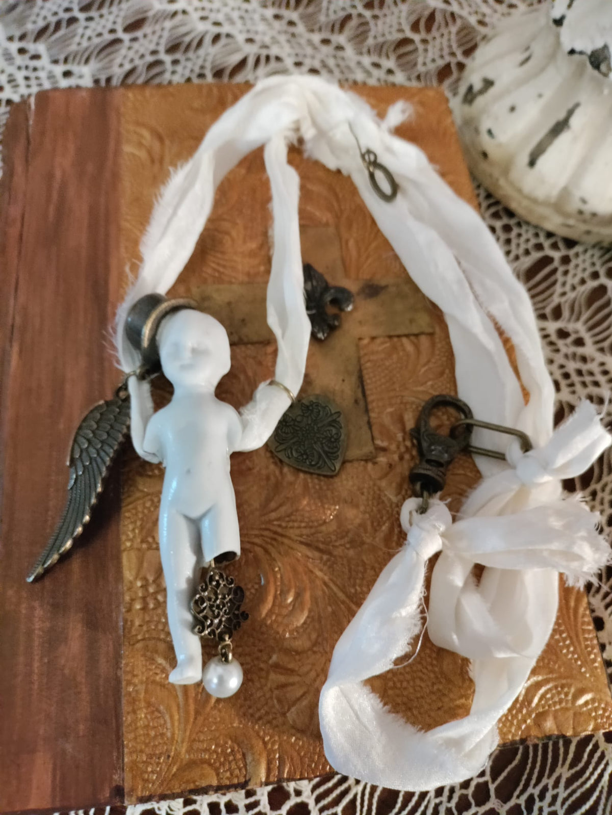 Cypher in the Snow pendant close-up on textured leather, bisque doll figure with bronze wing and heart charms among ivory silk ribbons