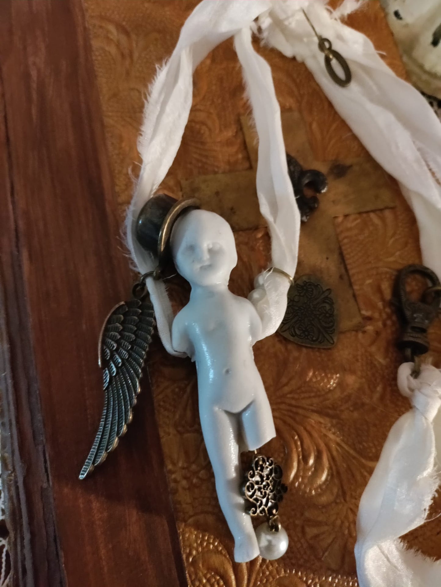 Close-up of Cypher in the Snow pendant, bisque doll wearing a small top hat with bronze angel wing, filigree heart charm, and pearl drop