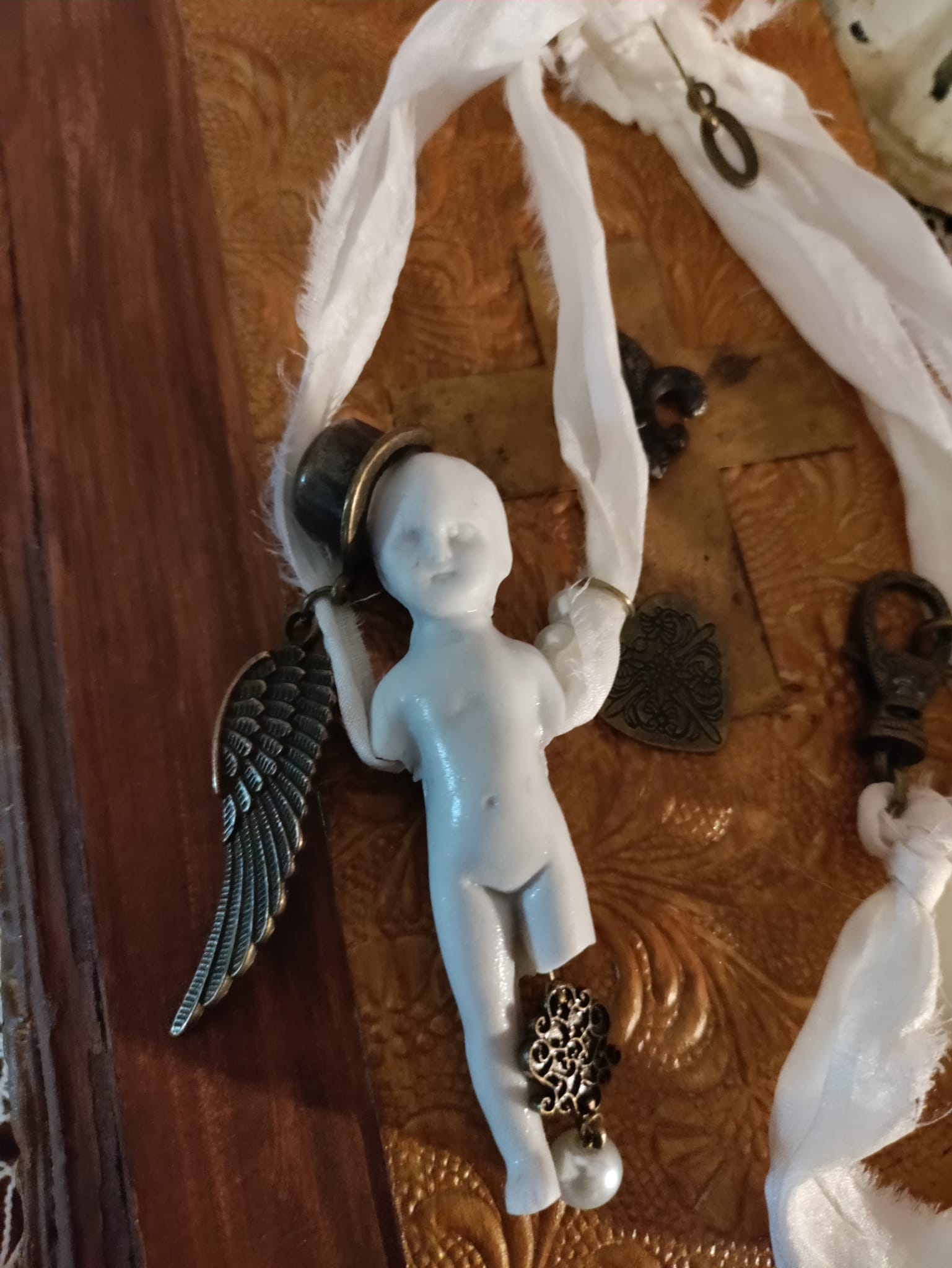 Close-up of Cypher in the Snow pendant, bisque doll wearing a small top hat with bronze angel wing, filigree heart charm, and pearl drop