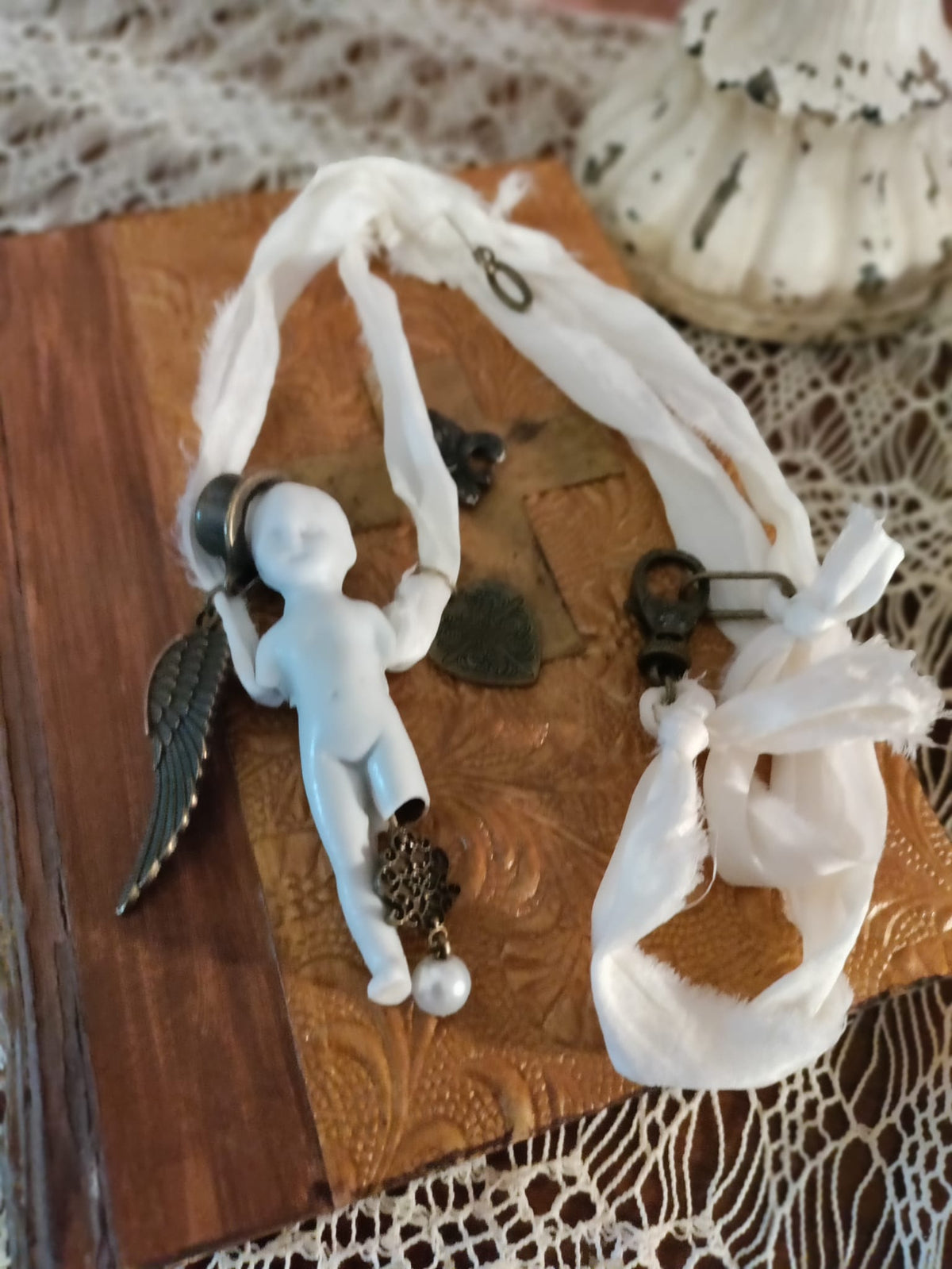 Cypher in the Snow necklace flat lay on an embossed leather book, bisque doll in a top hat with bronze wing and heart charms and a pearl drop