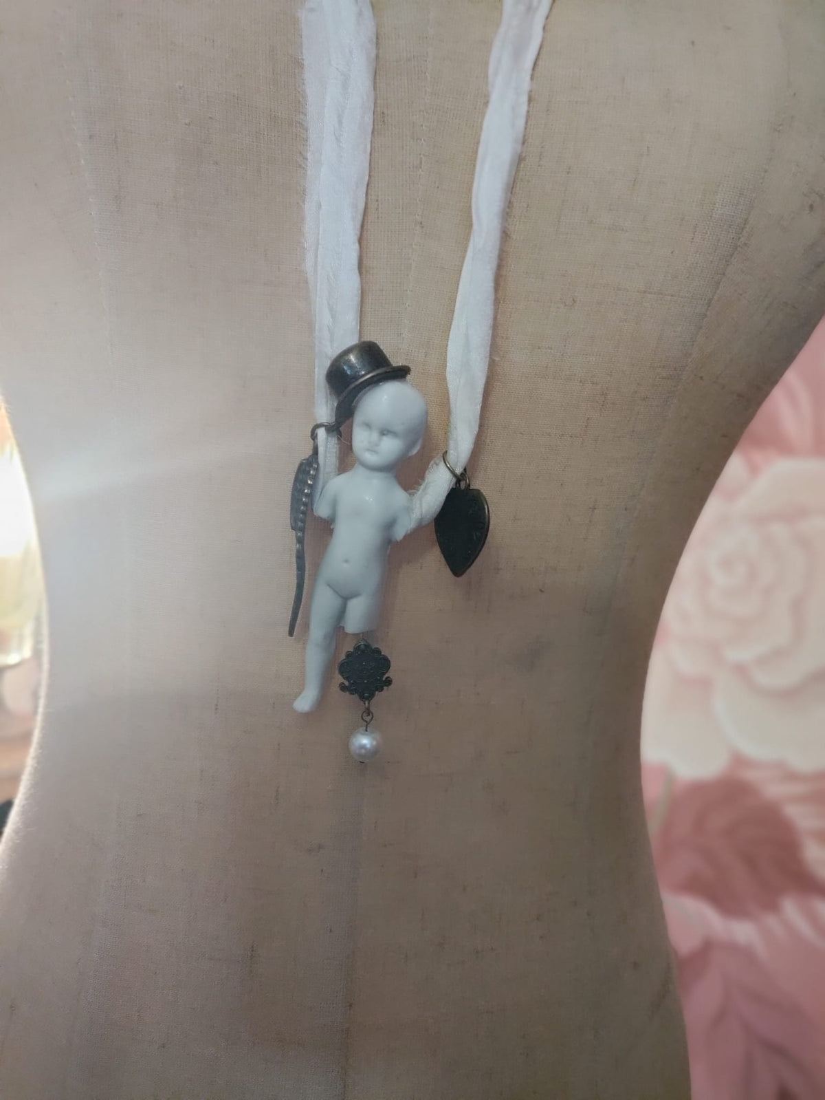 Close-up of Cypher in the Snow pendant on mannequin, bisque doll in a black top hat with bronze wing, heart charm, and pearl drop