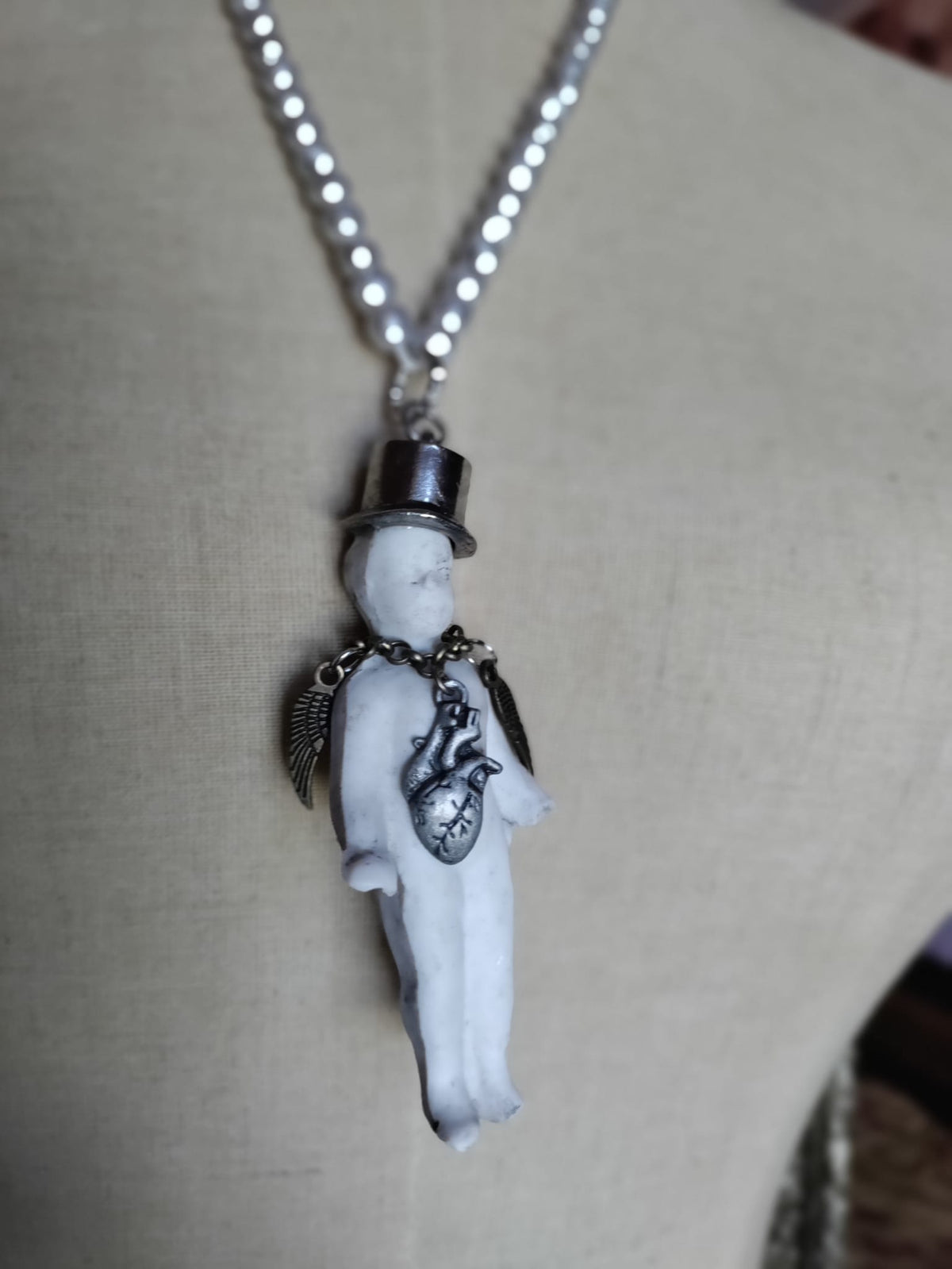 Close-up of Charlie's Broken Heart pendant on mannequin, bisque doll figure with silver top hat, angel wings, and pewter anatomical heart charm