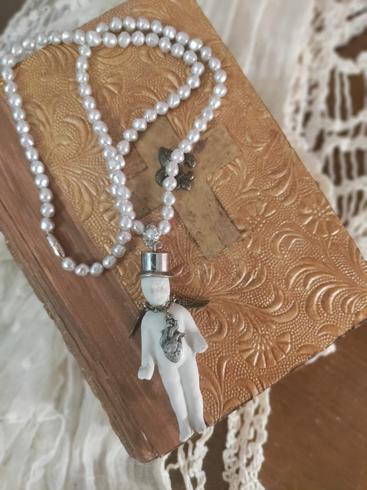 Charlie's Broken Heart necklace flat lay on an embossed leather book, pearl bead chain with bisque doll pendant in a silver top hat