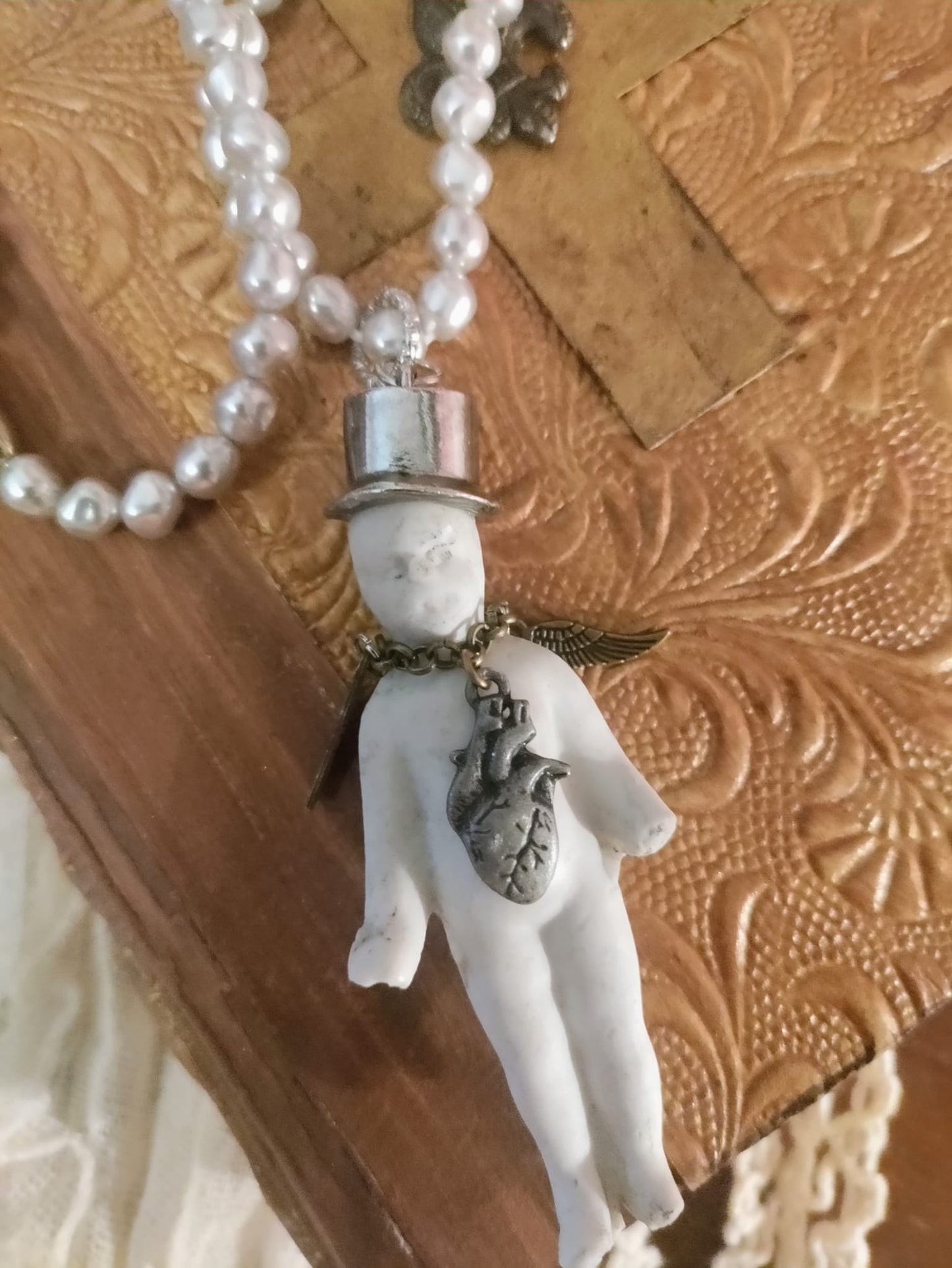 Charlie's Broken Heart pendant close-up, bisque doll in a silver top hat with bronze angel wing and pewter anatomical heart charm on a pearl bead chain, resting on an embossed leather book