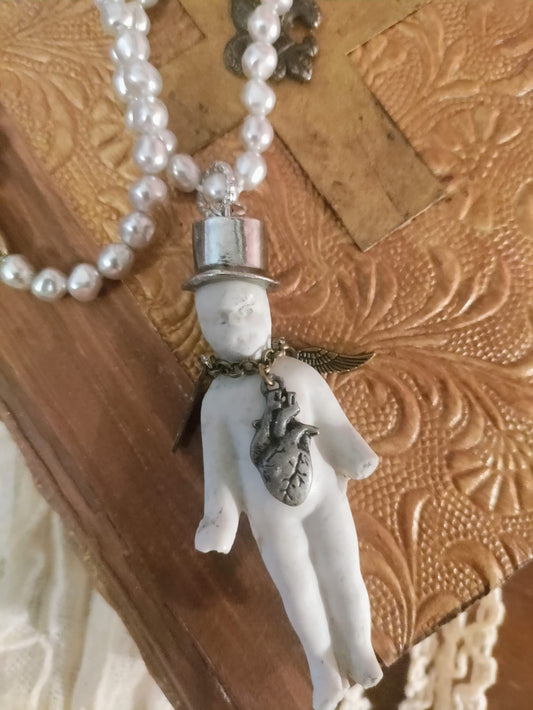 Charlie's Broken Heart pendant close-up, bisque doll in a silver top hat with bronze angel wing and pewter anatomical heart charm on a pearl bead chain, resting on an embossed leather book