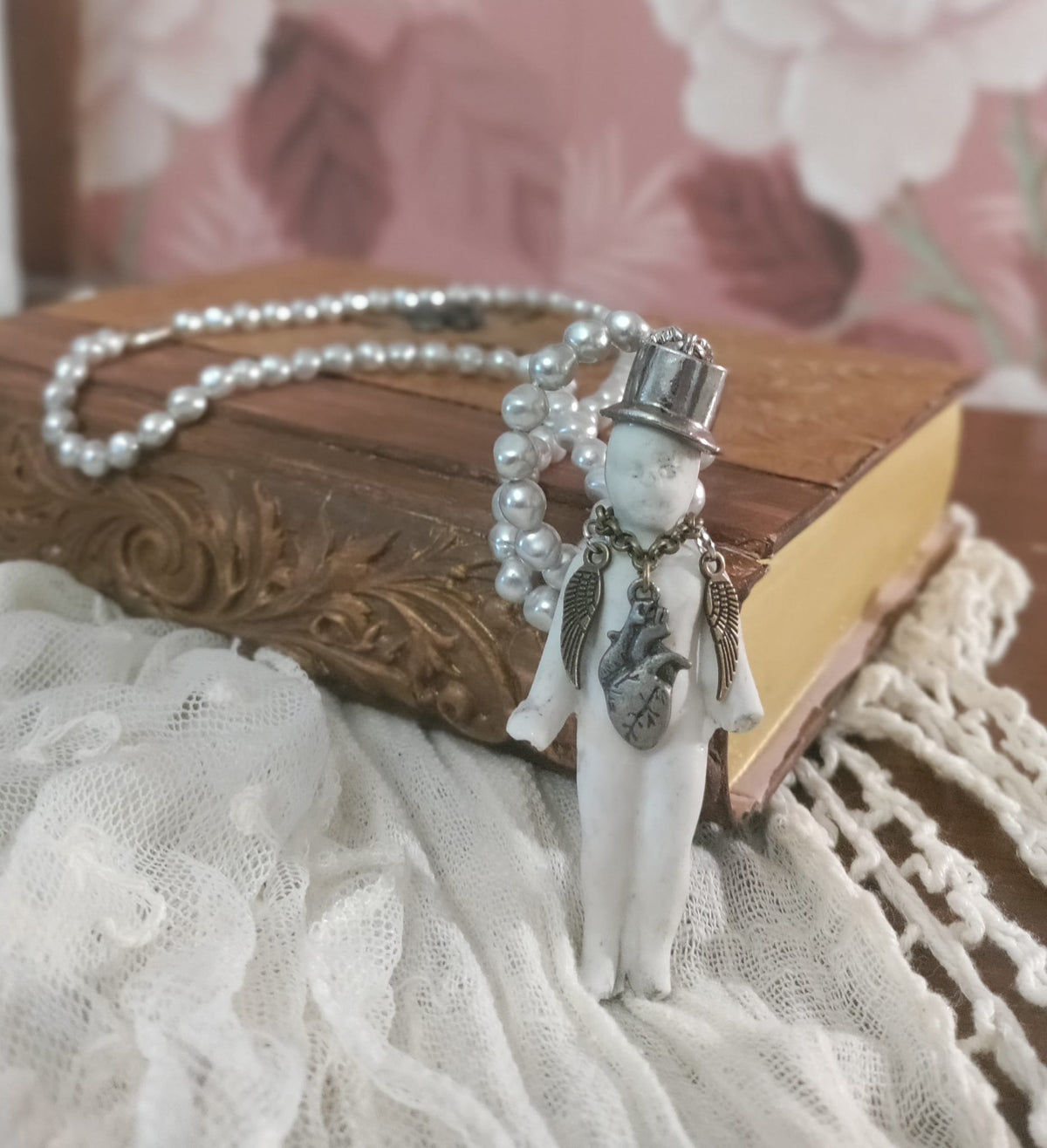 Charlie's Broken Heart necklace flat lay, pearl bead chain and bisque doll pendant with silver top hat and pewter heart charm on a book beside lace