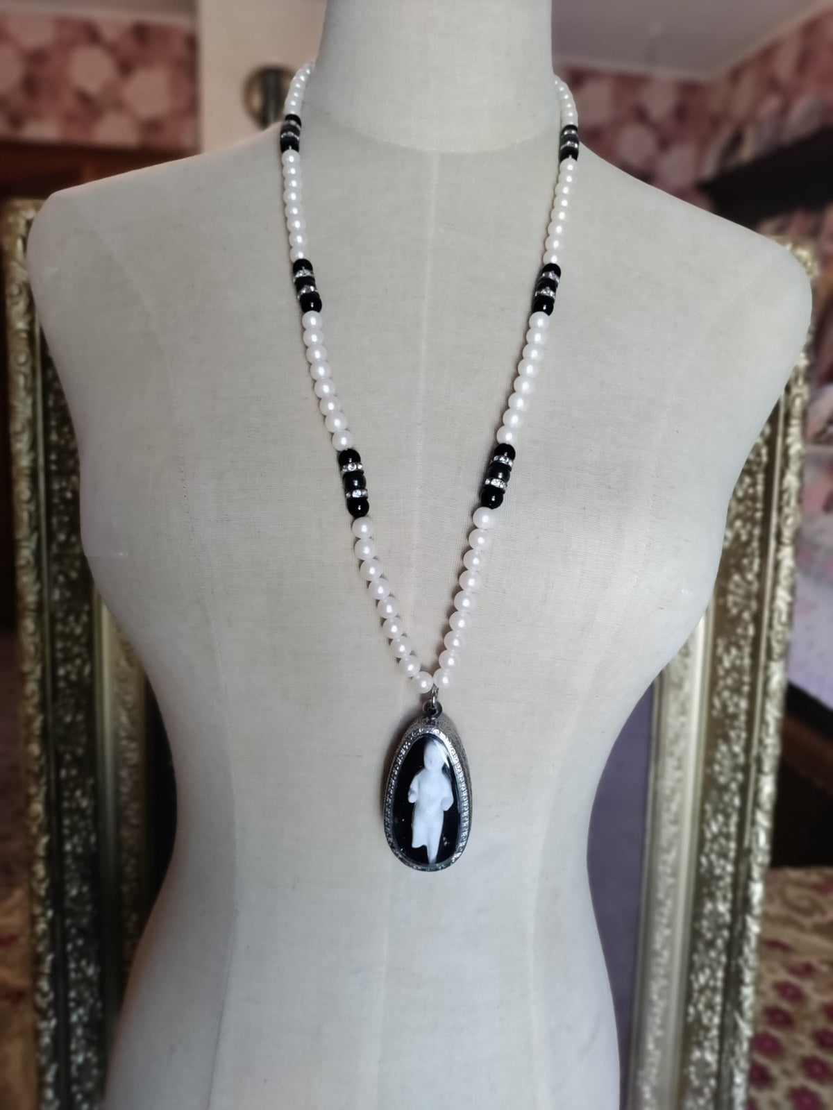 Reliquary Frozen Charles necklace on mannequin against framed mirrors, pearl and black bead chain with oval bisque doll pendant