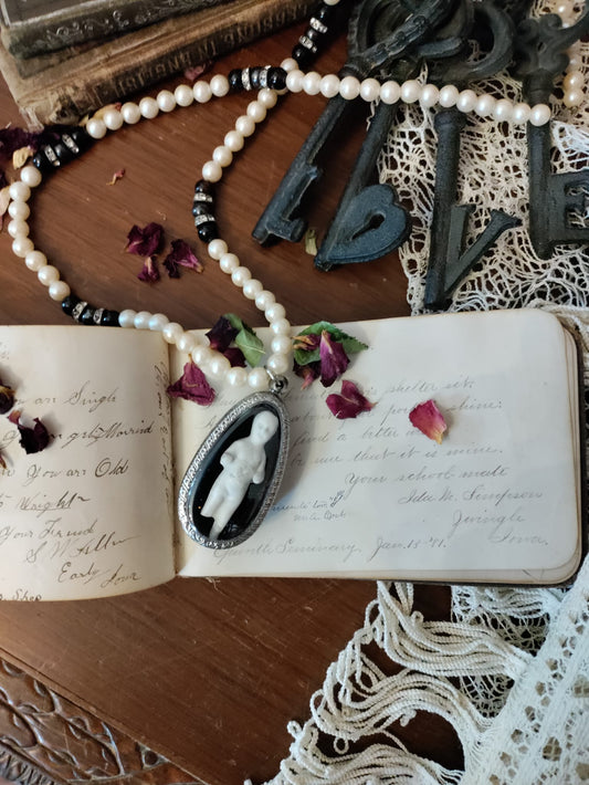 Reliquary Frozen Charles necklace flat lay, oval silver bezel pendant with antique bisque figure resting on an antique handwritten autograph book, surrounded by dried rose petals, iron skeleton keys, and lace