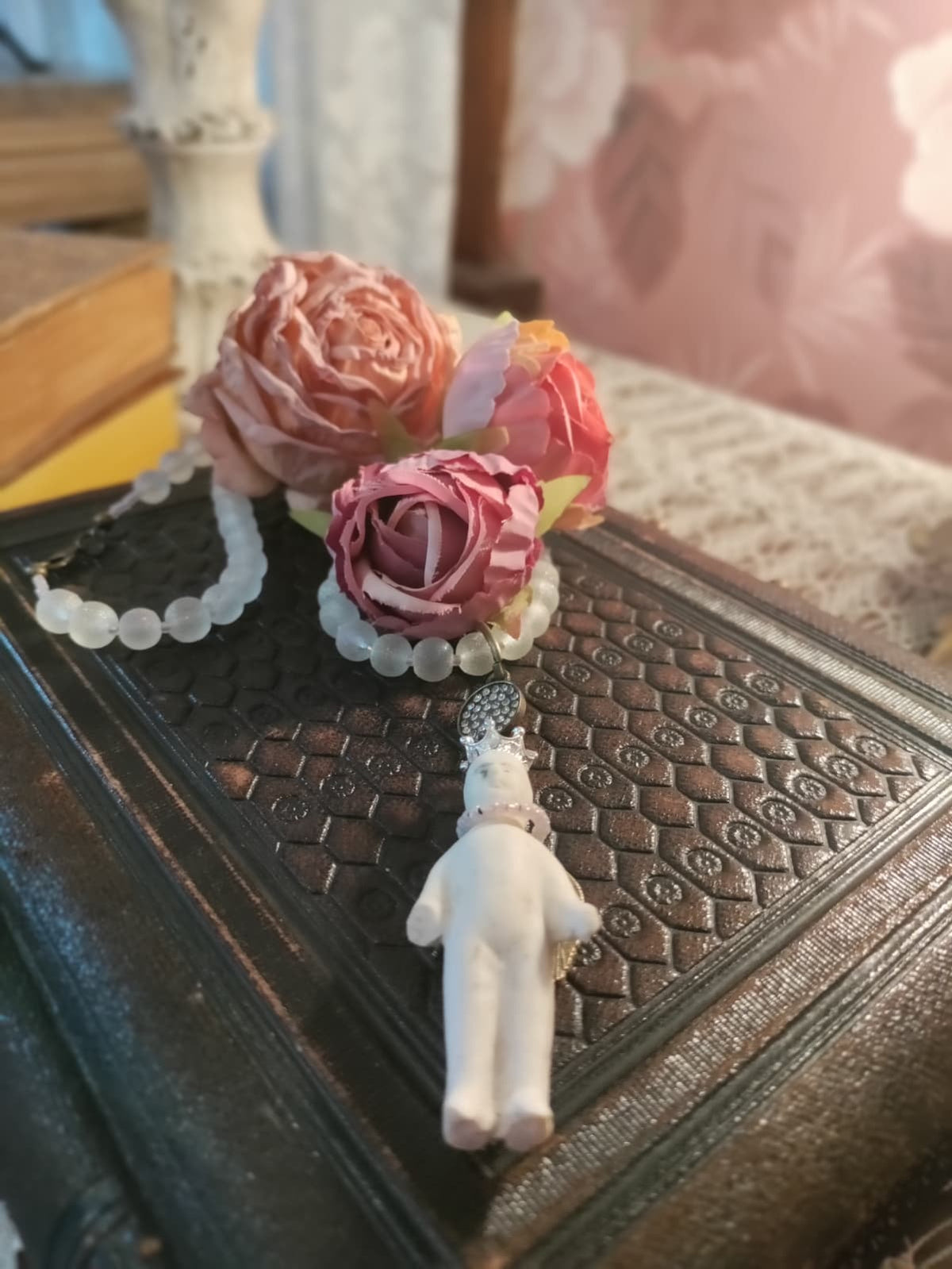 Frozen Charlotte necklace resting beside dried pink rose blooms on an antique book, bisque doll pendant with crown and rhinestone connector against floral wallpaper