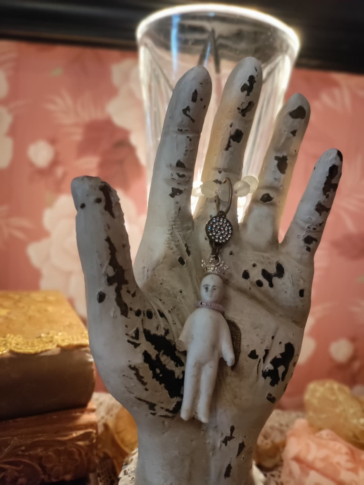 Frozen Charlotte necklace hanging from a weathered display hand, with an antique glass and stacked books in the background