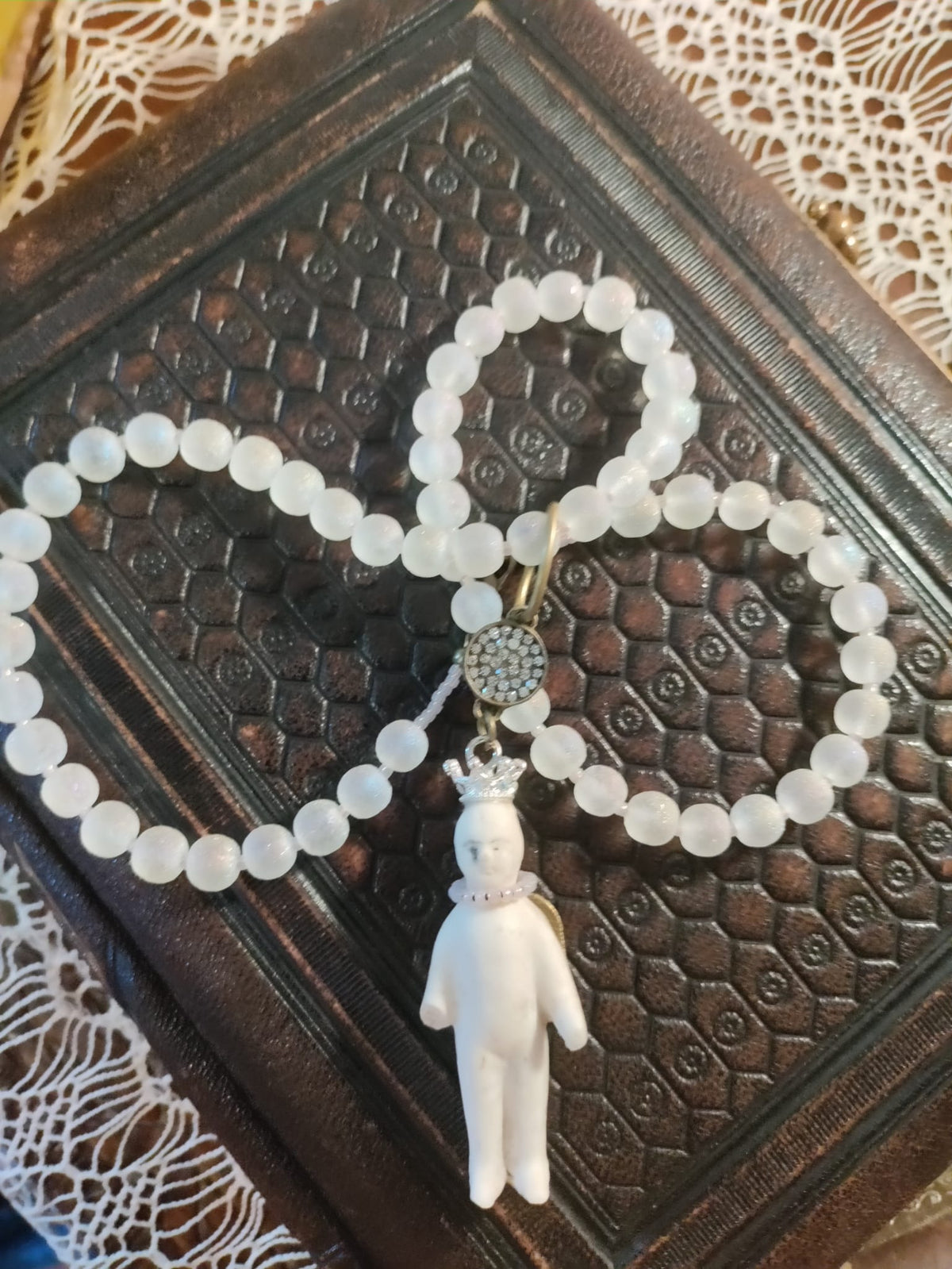 Frozen Charlotte necklace draped over an antique embossed leather book, frosted white glass beads coiled beside the crowned bisque doll pendant
