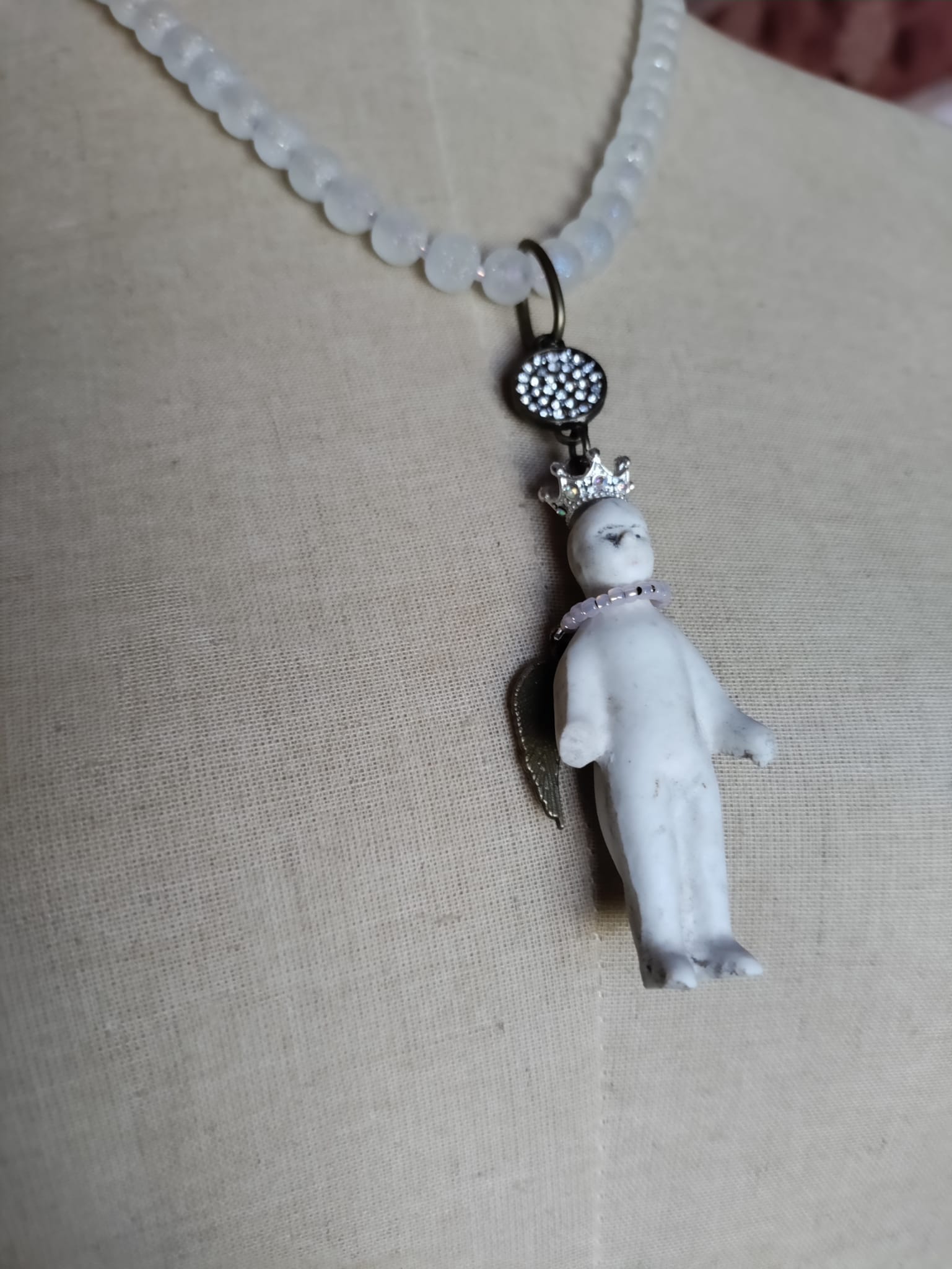 Close-up of the antique bisque Frozen Charlotte doll pendant wearing a silver rhinestone crown and pink seed bead collar, with a small bronze angel wing and rhinestone pavé connector above