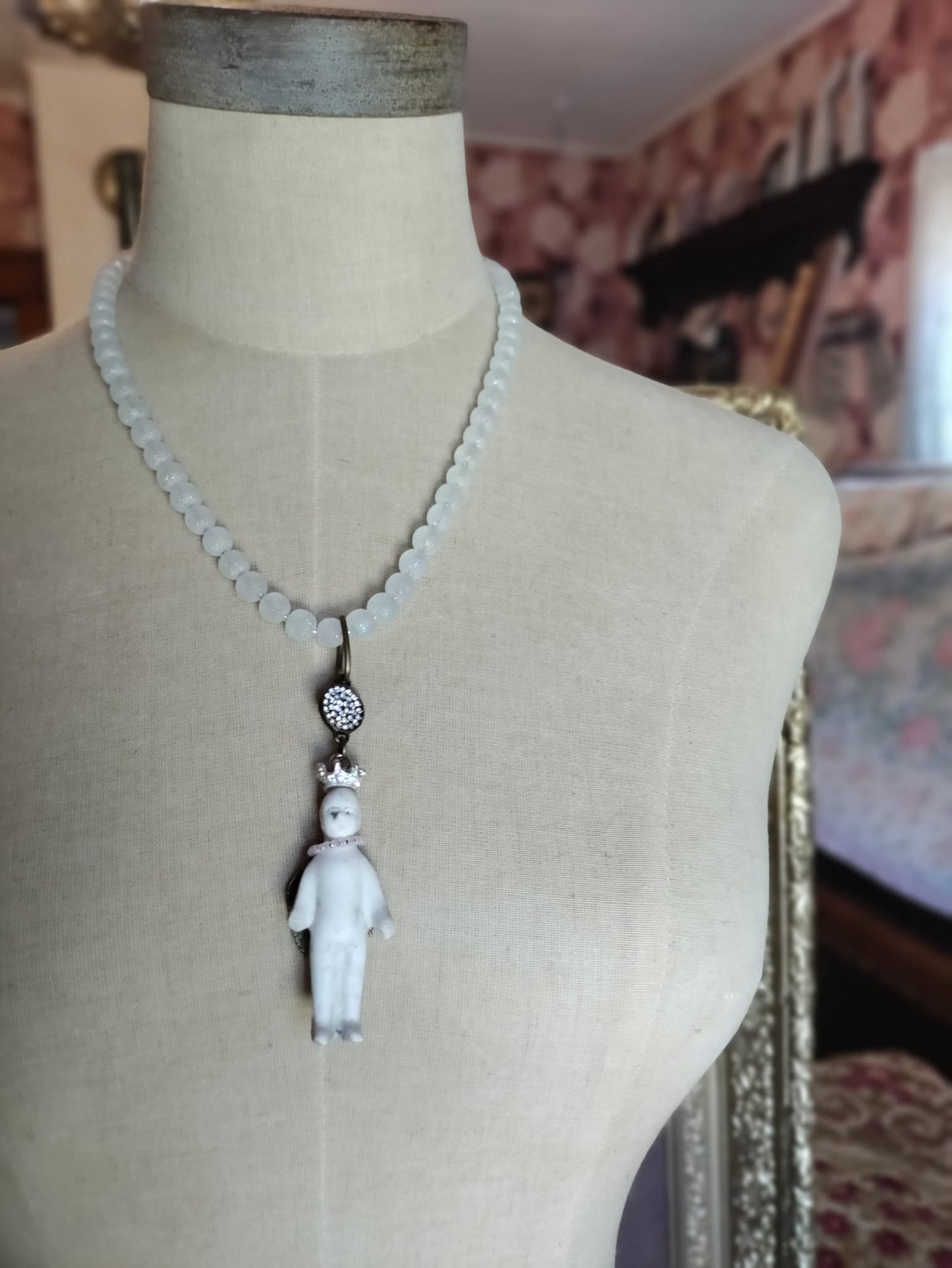 Frozen Charlotte necklace on a fabric dress form, showing the full frosted white glass bead strand and antique bisque doll pendant with silver rhinestone crown