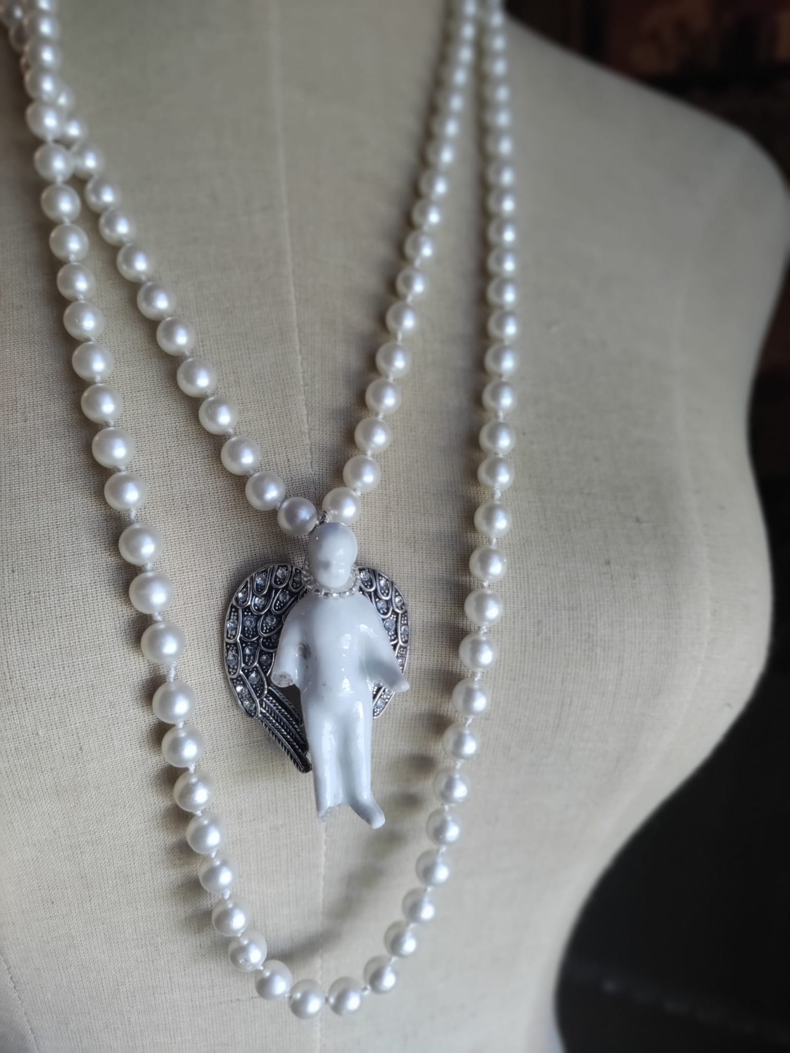 Frozen Charlotte necklace worn doubled on a dress form, showing the bisque doll pendant with silver rhinestone angel wings against the knotted pearl strands