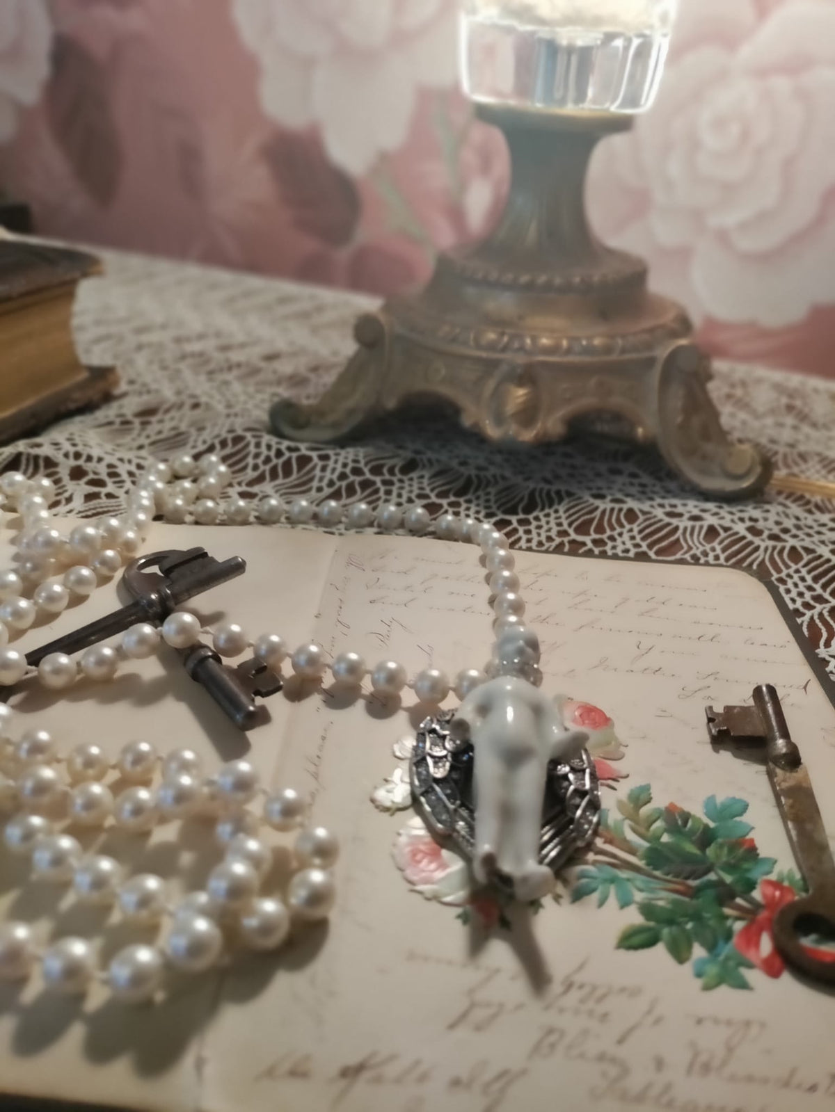 Frozen Charlotte necklace coiled on a lace-covered table with antique skeleton keys and a brass candlestick, pendant resting on a handwritten vintage letter