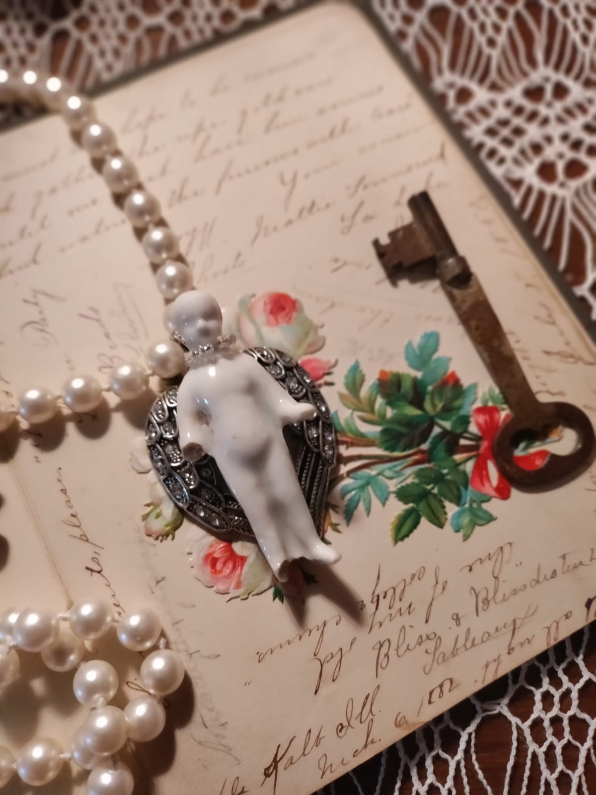 Frozen Charlotte pendant with silver rhinestone angel wings resting face-down on an antique handwritten letter beside a pressed floral card and skeleton key