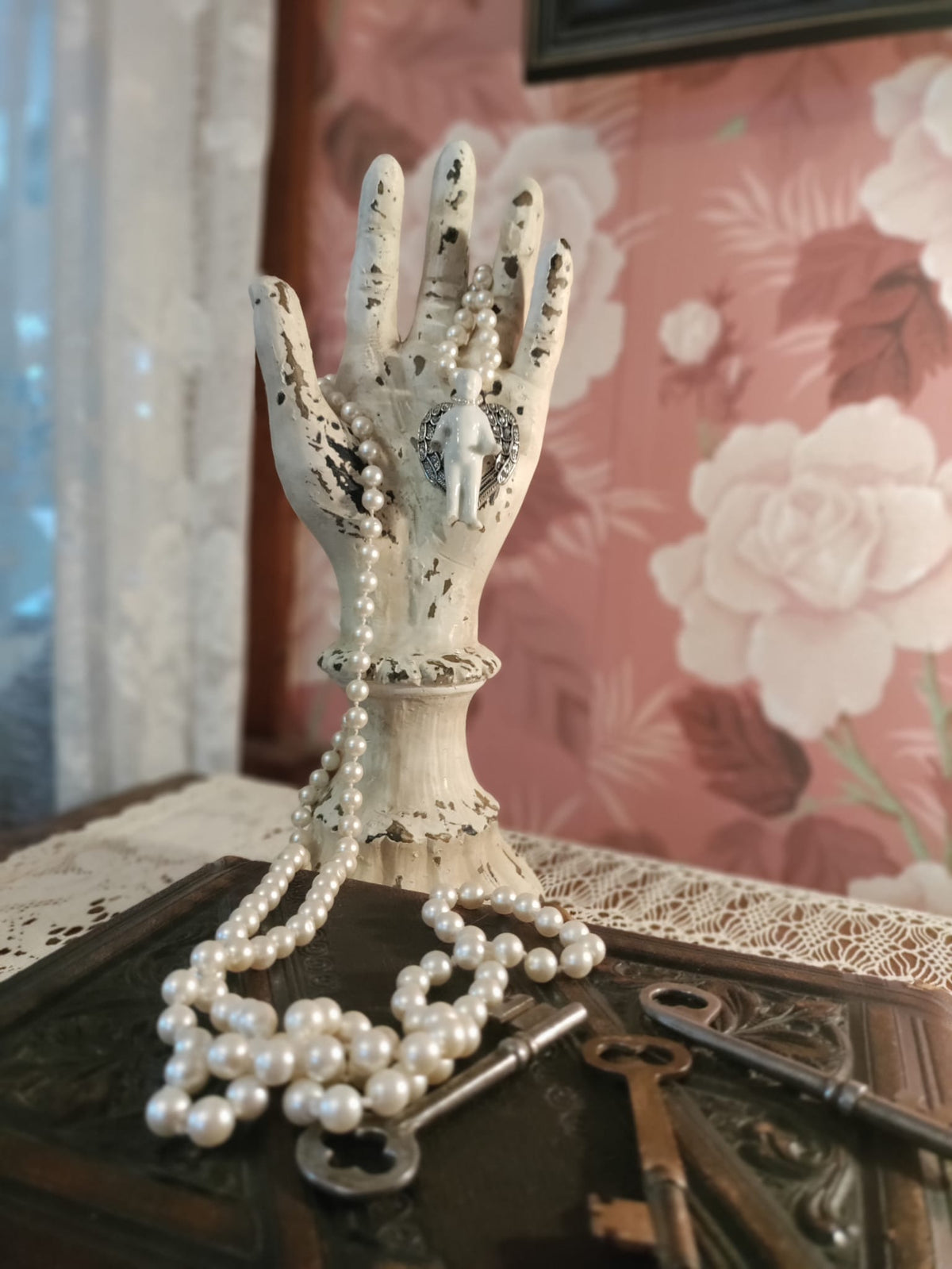 Frozen Charlotte necklace draped on a weathered display hand, long knotted strand of vintage 1920s glass pearls with the bisque doll and silver angel wing pendant, antique keys resting on a box below